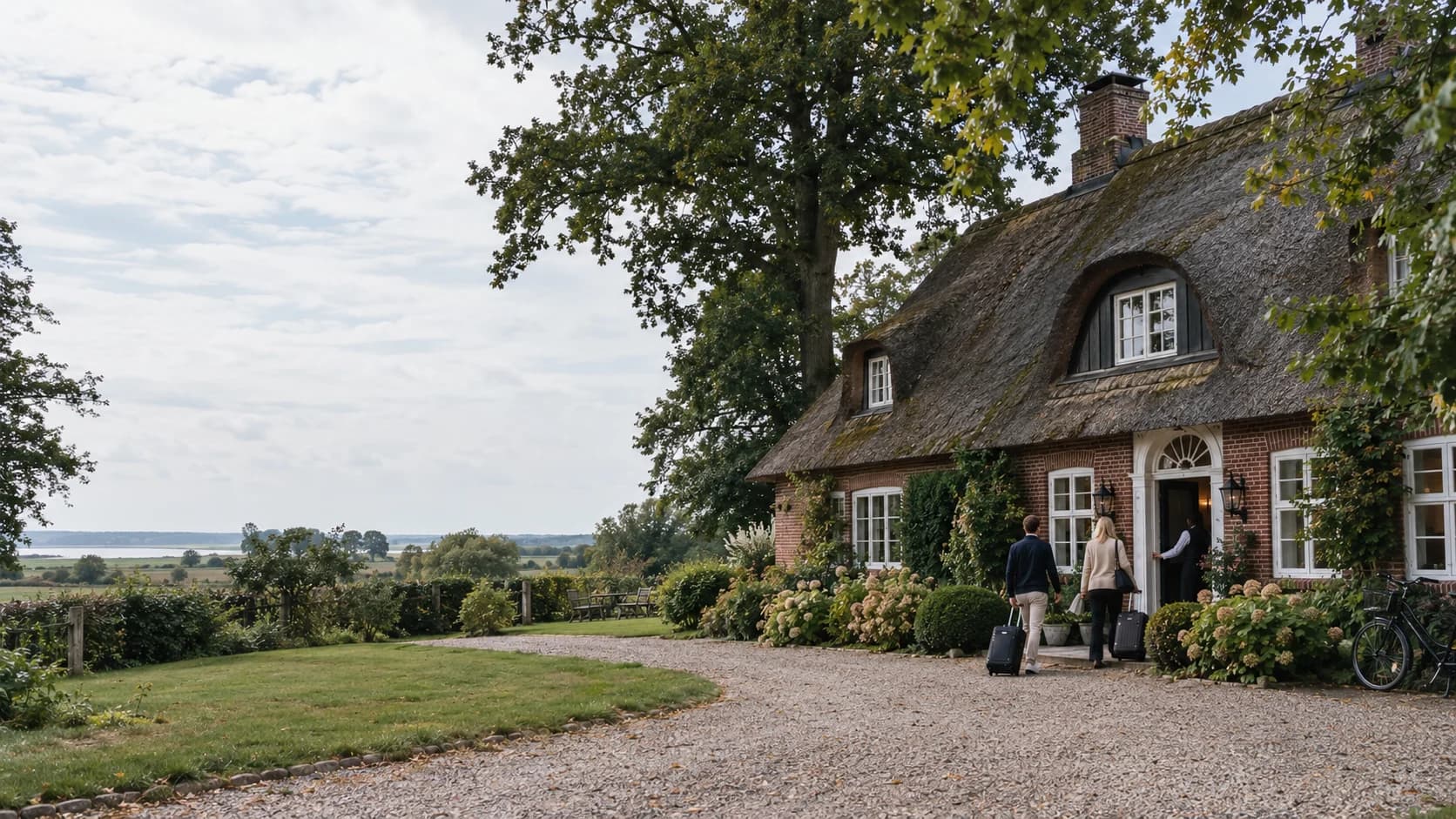 Symbolbild: Landhotel mit Reetdach und Backstein in Schleswig-Holstein, ein Paar mit Koffern wird an der Tür begrüßt (KI-generiert)